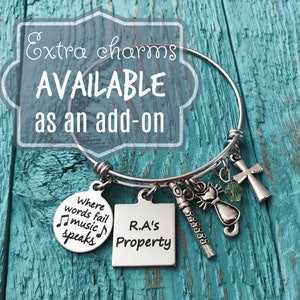 May include: A silver charm bracelet with various charms, including a music note charm with the text "Where words fail music speaks", a square charm with the text "R.A's Property", a cat charm, a cross charm, and a green crystal charm.