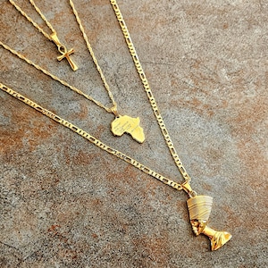 May include: A layered gold-tone necklace set featuring three distinct chains. One chain holds an ankh pendant, another features a map of Africa pendant, and the longest chain displays a detailed Nefertiti bust pendant with a textured crown.
