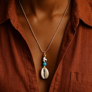 Può includere: Una collana color oro con un ciondolo a forma di conchiglia, una perla turchese, una perla bianca e nera e una perla a forma di pallone da calcio. La collana è indossata su una camicia marrone.