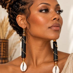 May include: Dangle earrings featuring a long matte black cylindrical bead wrapped in thin gold-tone wire, accented with a faceted amber-colored bead and a white cowrie shell at the base, suspended from a gold-tone hook with the text KHEPERA ADORNMENTS LLC.