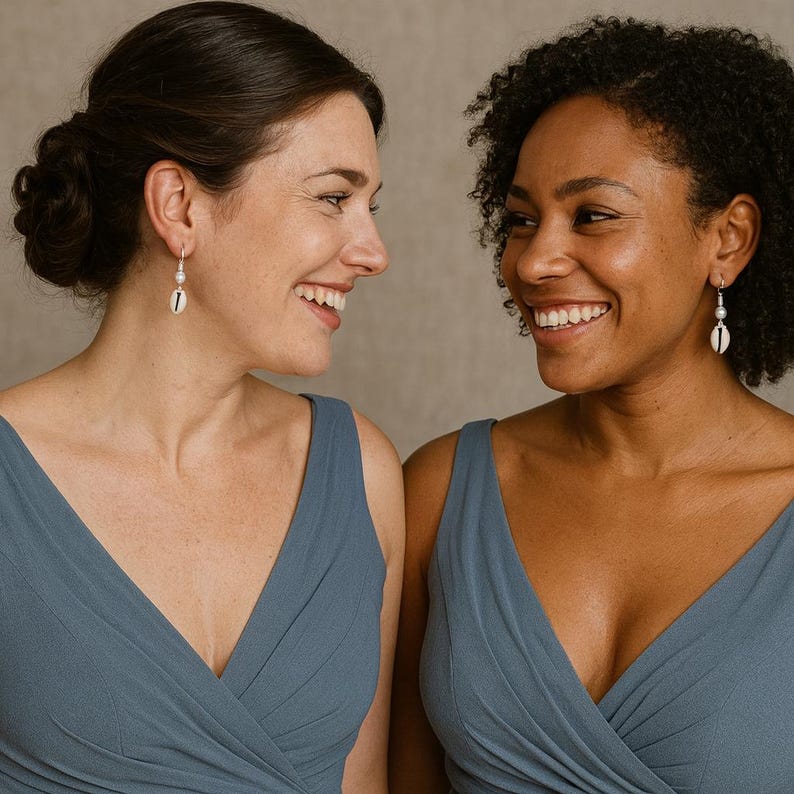 May include: Two women wearing matching blue dresses and shell earrings. The earrings feature a small shell pendant and a pearl. The women are smiling and looking at each other.