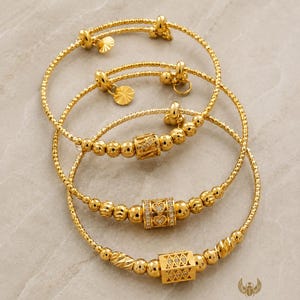Op de afbeelding: Drie goudkleurige armbanden met kralenkettingen en centrale bedels. Eén bedel is cilindervormig met hartvormige uitsparingen, een andere is een rechthoekig blok met diamantachtige accenten en de derde is een getextureerde cilinder.