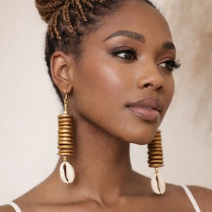 May include: Gold-coloured, stacked wooden earrings with a seashell accent. The earrings hang from a gold hook. The image also features the text "KHEPERA ADORNMENTS LLC".
