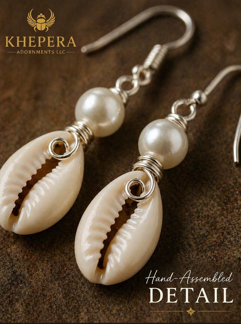 May include: A pair of handcrafted earrings featuring natural cowrie shells and faux pearls. The shells are cream-colored, and the pearls are white. The earrings are accented with silver-colored wire and hooks. The text "Hand-Assembled DETAIL" is visible.