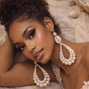 May include: A woman wearing large, teardrop-shaped pearl earrings. The earrings feature small pearls and a seashell accent. The woman has dark hair and is wearing makeup. The earrings are a statement piece.