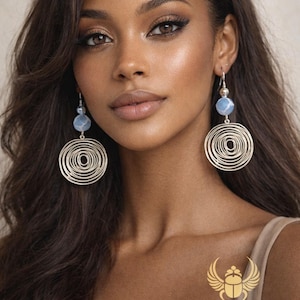 May include: Silver spiral earrings with a blue bead detail. The earrings hang from silver hooks. The earrings are part of the KHEPERA ADORNMENTS LLC collection.