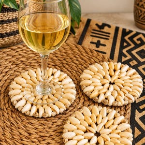 Puede incluir: Tres posavasos circulares hechos de pequeñas conchas ovaladas de color claro dispuestas en un patrón radial, con un borde texturizado de diminutas cuentas color canela. Un posavasos sostiene un vaso con una bebida de color amarillo pálido.