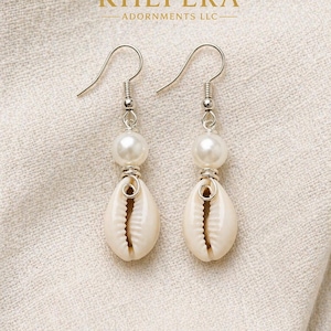 May include: A pair of dangle earrings featuring a white pearl bead above a natural seashell. The earrings have silver-colored hooks and accents. The text "KHEPERA ADORNMENTS LLC" is visible at the top. The text "Lightweight &bull; Comfortable &bull; Easy to Wear" is at the bottom.