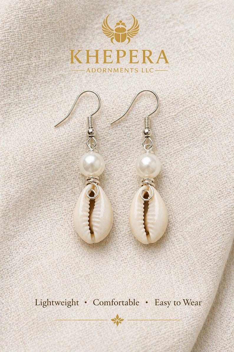 May include: A pair of dangle earrings featuring a white pearl bead above a natural seashell. The earrings have silver-colored hooks and accents. The text "KHEPERA ADORNMENTS LLC" is visible at the top. The text "Lightweight &bull; Comfortable &bull; Easy to Wear" is at the bottom.