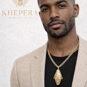 May include: A gold-colored pendant necklace with a stylized face design hangs from a thick chain. The model is wearing a black shirt and a tan blazer. The brand name "KHEPERA ADORNMENTS LLC" is visible.