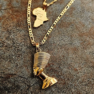 May include: A layered gold-tone necklace set featuring three distinct pendants: a polished Nefertiti bust, a textured map of the African continent, and a classic Ankh cross symbol. Each pendant hangs from a matching gold-tone chain of varying lengths.