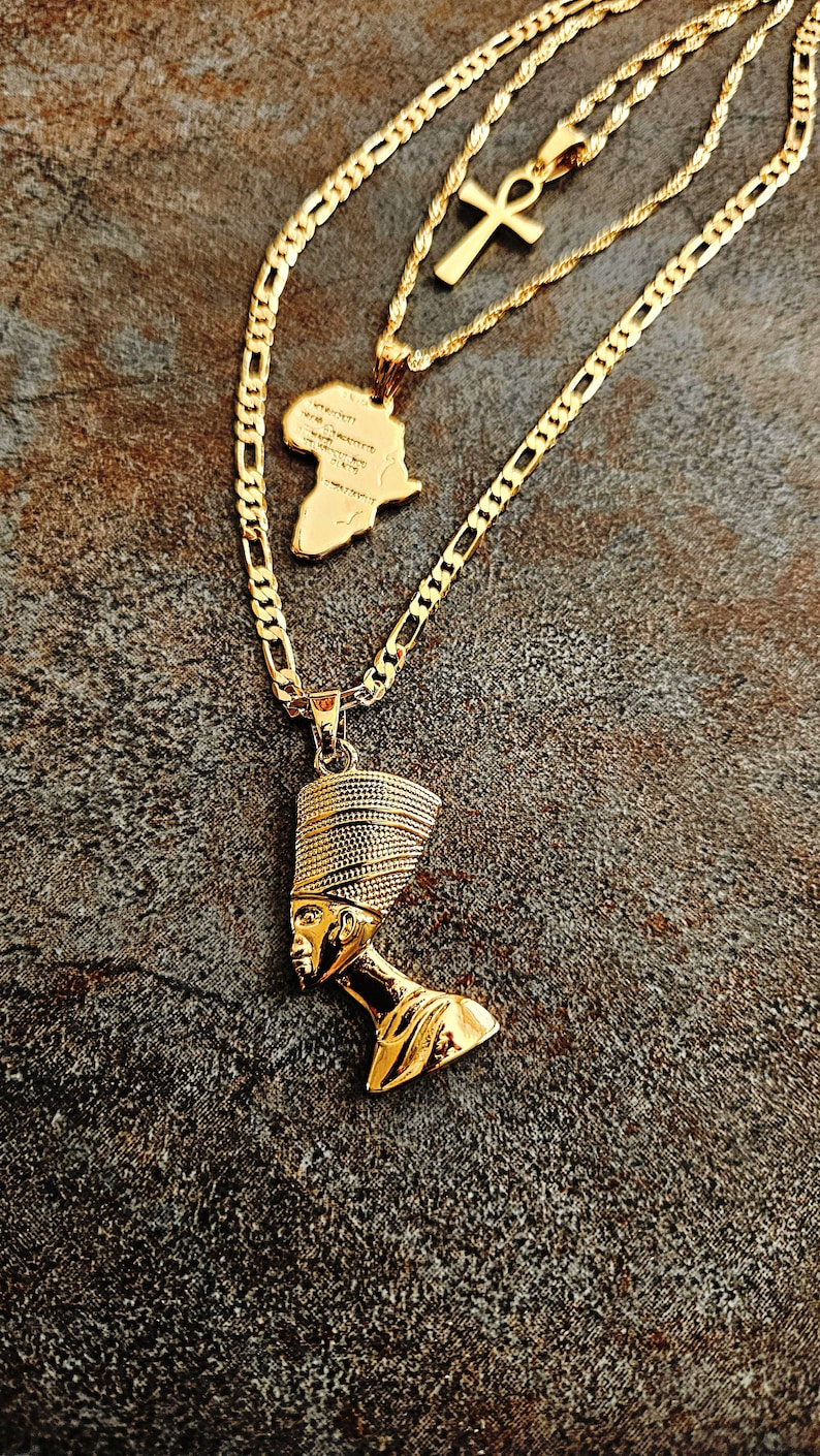 May include: A layered gold-tone necklace set featuring three distinct pendants: a polished Nefertiti bust, a textured map of the African continent, and a classic Ankh cross symbol. Each pendant hangs from a matching gold-tone chain of varying lengths.