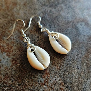 May include: A pair of silver wire earrings with white cowrie shell pendants. The shells are attached to the wire with a silver wire wrap.