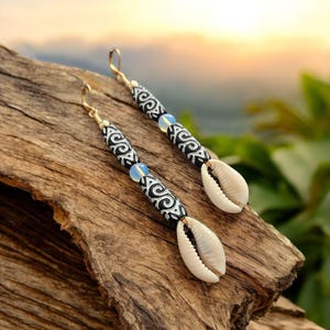 May include: A pair of dangle earrings featuring white cowrie shells, black and white patterned beads, and gold-colored hooks. The earrings also have small, light blue beads. The earrings are displayed on a piece of wood.
