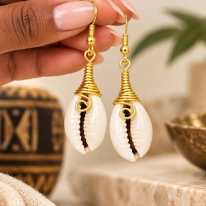 May include: A pair of dangle earrings featuring white cowrie shells wrapped in gold-toned wire. The shells are suspended from gold-toned fishhook ear wires. The design includes a coiled wire detail at the top of each shell, creating a drop-style earring.