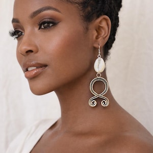 May include: A woman wearing long, dangling earrings. The earrings have a white seashell at the top, connected to a gold-coloured, ornate design. The earrings are part of the KHEPERA ADORNMENTS LLC collection.