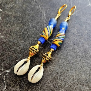 Puede incluir: Un par de aretes largos de oro con cuentas de vidrio azul y amarillo y detalles de concha blanca.