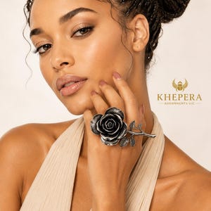 May include: A large silver-toned ring featuring a detailed three-dimensional rose bloom with a stem and leaves extending across the finger. The metal has an oxidized finish to highlight the texture of the petals and foliage. The logo KHEPERA ADORNMENTS LLC is visible.