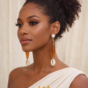 May include: Long, dangling earrings with a pearl top, amber-colored center, and a white seashell pendant. The earrings are worn by a person wearing a white, off-the-shoulder top. The brand name "KHEPERA ADORNMENTS LLC" is visible.