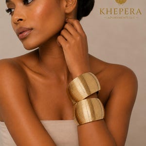 May include: Two gold-colored, wide cuff bracelets with a ribbed design. The bracelets are worn on the wrist. The image also includes the text "KHEPERA - ADORNMENTS LLC -" and a scarab beetle logo.