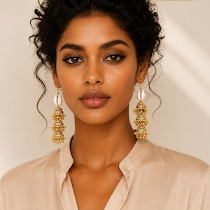 May include: Gold-toned chandelier earrings with a tiered bell design and small bead accents. The earrings feature a white shell at the top. The earrings are worn by a person wearing a beige collared shirt. The logo "KHEPERA - ADORNMENTS LLC" is visible.