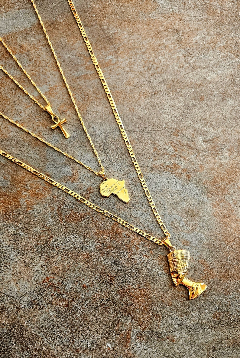 May include: A layered gold-tone necklace set featuring three distinct chains. One chain holds an ankh pendant, another features a map of Africa pendant, and the longest chain displays a detailed Nefertiti bust pendant with a textured crown.