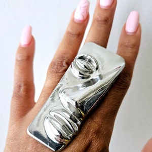 May include: A silver-colored ring with a rectangular shape and a raised, pill-like design. The ring is worn on a hand with pink nail polish.
