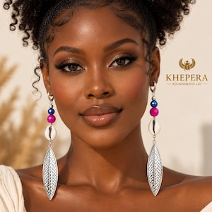 May include: Dangle earrings featuring a silver-toned hoop, a faceted blue bead, a faceted magenta bead, a white cowrie shell, and a long, pointed silver-toned leaf-shaped charm with a textured herringbone pattern. The text KHEPERA ADORNMENTS LLC is visible.