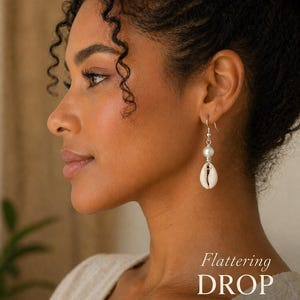 May include: Drop earrings featuring a white pearl and a cowrie shell pendant. The earrings are suspended from a silver hook. The image includes the text "Flattering DROP LENGTH" and the brand name "KHEPERA - ADORNMENTS LLC".