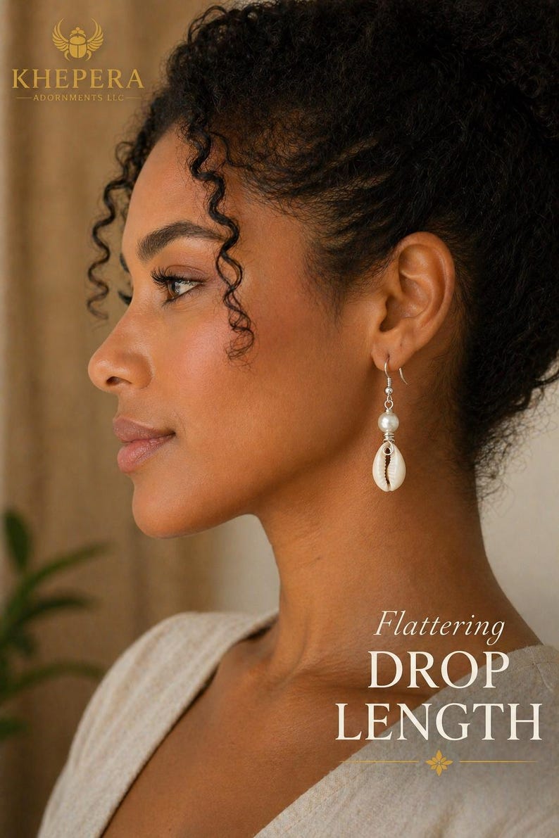 May include: Drop earrings featuring a white pearl and a cowrie shell pendant. The earrings are suspended from a silver hook. The image includes the text "Flattering DROP LENGTH" and the brand name "KHEPERA - ADORNMENTS LLC".