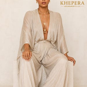 May include: A champagne-coloured, pleated jumpsuit featuring a deep V-neckline, wide-leg trousers, and flowing kimono-style sleeves. The garment includes a thin waist tie and is paired with gold-toned layered necklaces and small gold earrings.
