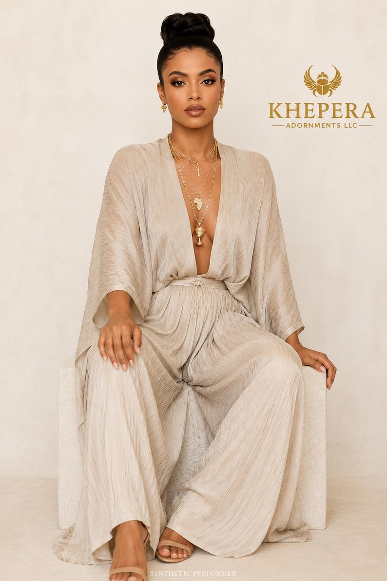 May include: A champagne-coloured, pleated jumpsuit featuring a deep V-neckline, wide-leg trousers, and flowing kimono-style sleeves. The garment includes a thin waist tie and is paired with gold-toned layered necklaces and small gold earrings.