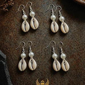 May include: Four pairs of earrings with a natural seashell and pearl design. Each earring features a cream-coloured shell, a white pearl, and silver-coloured wire. The earrings are displayed on a dark brown surface. The logo "KHEPERA ADORNMENTS LLC" is at the bottom.