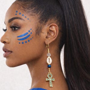 May include: A woman with dark hair in a high ponytail wearing gold earrings with a seashell, blue bead, and an Ankh pendant. Her face has blue face paint. She is wearing a blue top.