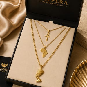 May include: A set of three gold-toned necklaces displayed in a black box labelled KHEPERA ADORNMENTS LLC. The necklaces feature an ankh cross, a map of Africa, and a Nefertiti bust pendant, all suspended on delicate gold-toned link chains.