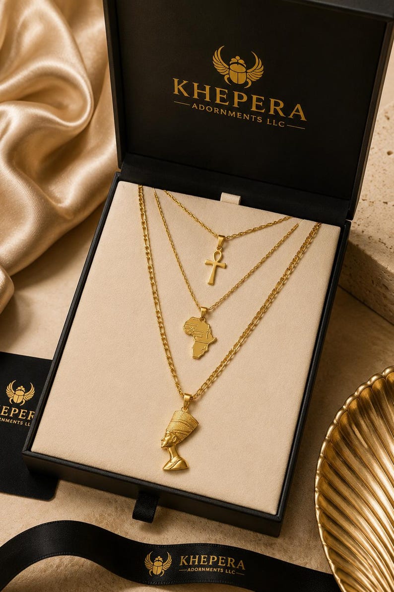 May include: A set of three gold-toned necklaces displayed in a black box labelled KHEPERA ADORNMENTS LLC. The necklaces feature an ankh cross, a map of Africa, and a Nefertiti bust pendant, all suspended on delicate gold-toned link chains.