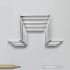 May include: A set of silver-colored, L-shaped metal tools arranged in a nested pattern, resembling a musical note. A pencil and a scale with inches and centimeters are in the foreground. The background is a light gray.