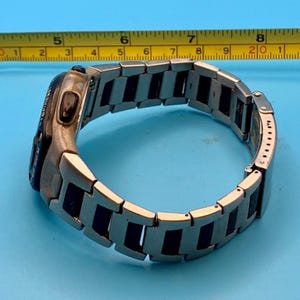May include: A silver and black metal link watch with a digital display, positioned next to a yellow measuring tape that shows the watch band has a diameter of approximately 2.5 inches. The watch features a side button labeled STOP/RESET.