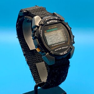May include: A black Armitron digital watch with a textured nylon strap. The watch face features the text "INSTALITE," "Armitron," "ALL-SPORT," "ALARM/CHRONO," "WATER RESIST," and "165 WR." It includes buttons labeled "MODE," "DATE," "ALARM," and "LIGHT."