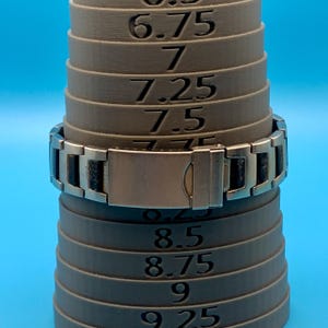 May include: A silver-toned metal link bracelet with black rectangular inlays, featuring a fold-over clasp. The bracelet is positioned on a grey tapered ring mandrel marked with numerical size increments ranging from 6.75 to 9.25 for measurement.