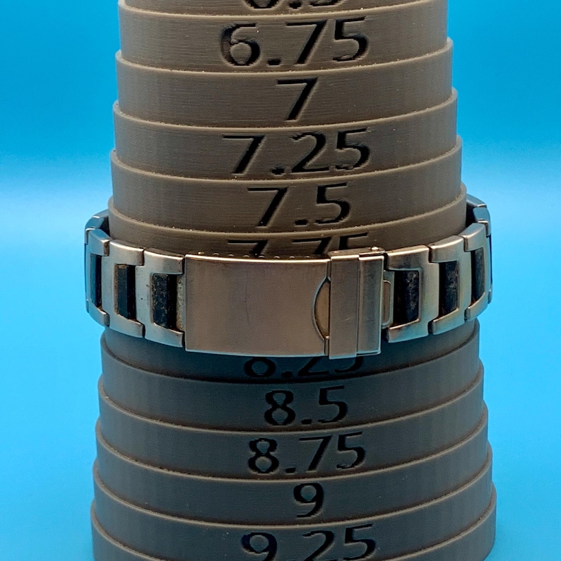 May include: A silver-toned metal link bracelet with black rectangular inlays, featuring a fold-over clasp. The bracelet is positioned on a grey tapered ring mandrel marked with numerical size increments ranging from 6.75 to 9.25 for measurement.