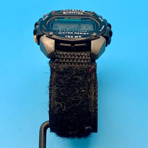 May include: A black Armitron All-Sport digital watch with a textured black fabric strap featuring a hook-and-loop closure. The watch face displays the text Armitron, All-Sport, Alarm Chrono, Water Resist, and 165 WR, with a yellow side button.