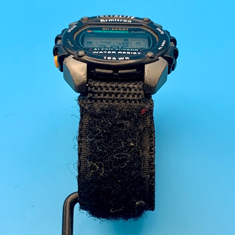 May include: A black Armitron All-Sport digital watch with a textured black fabric strap featuring a hook-and-loop closure. The watch face displays the text Armitron, All-Sport, Alarm Chrono, Water Resist, and 165 WR, with a yellow side button.