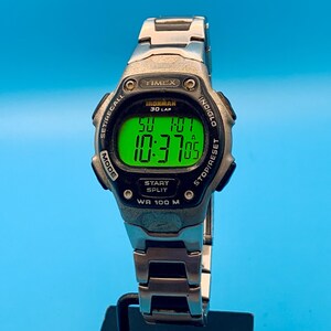 May include: A silver-tone Timex Ironman 30 Lap digital watch with a black bezel and a bright green backlit display. The screen shows the time 10:37:05 and the date SU 1-07. The watch features a metal link band and is marked WR 100 M on the face.