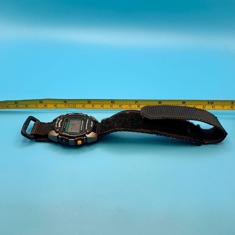 May include: A black digital Armitron watch with a fabric strap and velcro closure, positioned next to a yellow measuring tape that shows the watch and strap length extending to approximately 10.5 inches in total length.