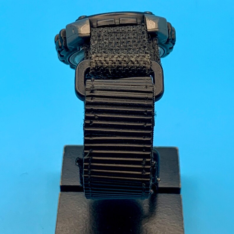 May include: A black digital wristwatch featuring a textured, ribbed black strap with a rectangular plastic buckle. The watch case is dark gray and black, showing a rugged design with visible side buttons and a woven fabric attachment point.