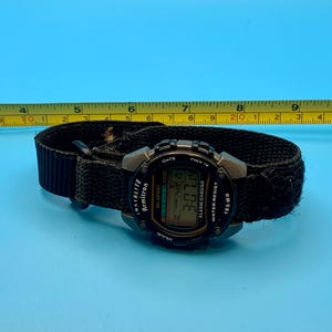 May include: A black digital Armitron All-Sport watch with a fabric strap, positioned next to a yellow measuring tape showing a length of approximately 9.5 inches. The watch face displays the time 3:07 and features text including Indiglo, Date, and 165 WR.
