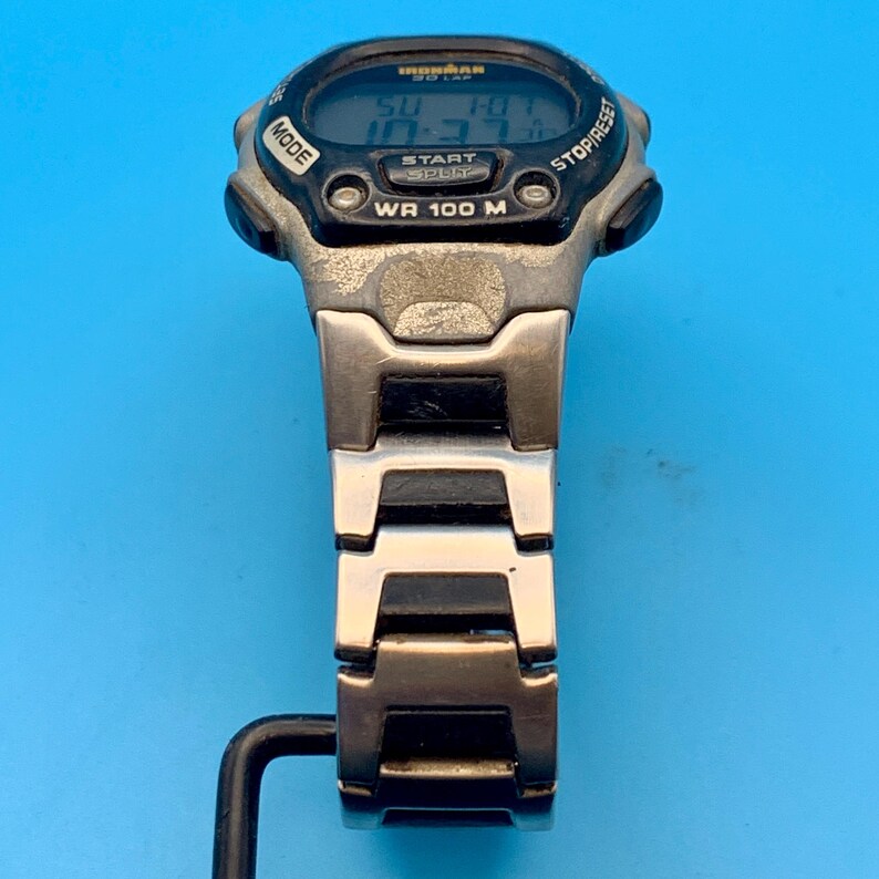 May include: A digital wristwatch featuring a black and silver-tone case with a digital display showing the text Ironman 30 Lap and WR 100 M. The watch has a metallic link bracelet with black accents and buttons labeled Mode and Stop/Reset.