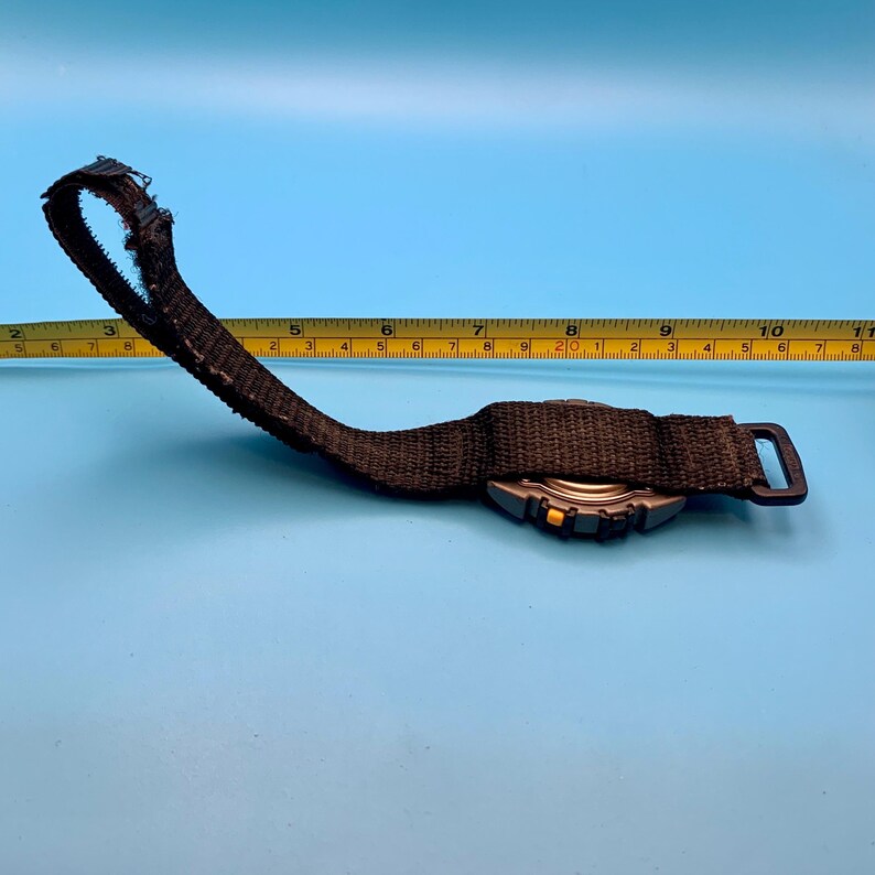May include: A black fabric watch strap with a dark grey watch case featuring a small yellow button, positioned next to a yellow measuring tape that shows the strap length extending past the 10-inch mark on the imperial scale for reference.