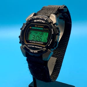 May include: A black digital Armitron All-Sport watch with a textured fabric strap. The watch face features a green digital display showing the time 3:06:12, with text including Indiglo, Alarm/Chrono, Water Resist, and 165 WR printed on the bezel.